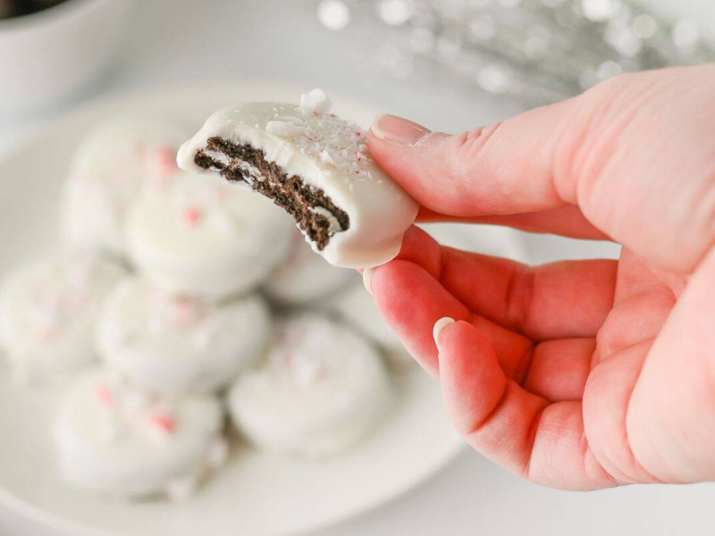 A hand holds a white chocolate-covered cookie with a bite taken out, while more similar cookies are visible on a plate in the background.