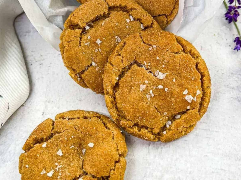 Three crackled ginger cookies topped with flakes of sea salt are arranged on a white surface with a piece of parchment paper and some purple flowers nearby.