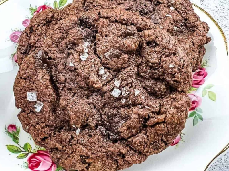 Two large, chocolate cookies topped with sea salt flakes are placed on a floral-patterned plate.