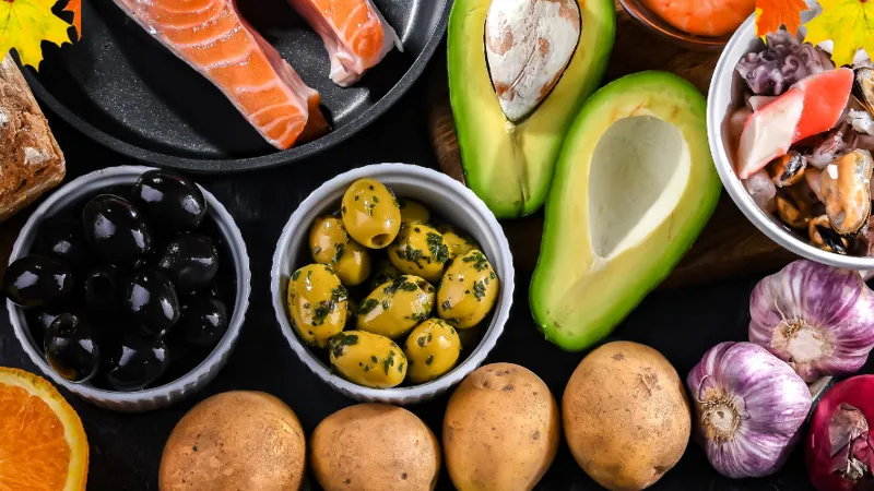 Add flavour to your reading week with these healthy, budget friendly recipes Assorted fresh fruits, vegetables, bread, salmon, olives, nuts, and shellfish arranged on a dark surface with autumn leaves scattered on top.