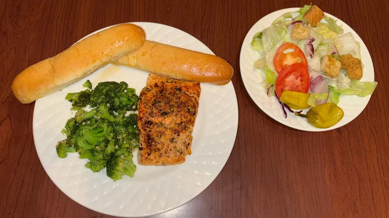Plate with salmon, broccoli and breadsticks and plate with salad on wood table.