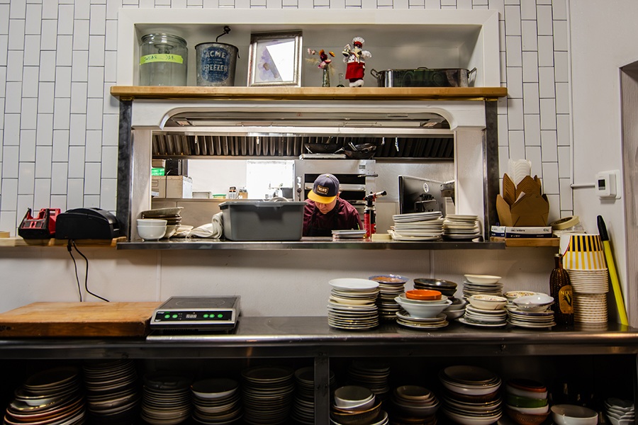 A cook in a baseball cap works in a restaurant kitchen, visible behind the pass. In front, stacks of mismatched plates fill shelves, and knickknacks including a swear jar decorate the space.