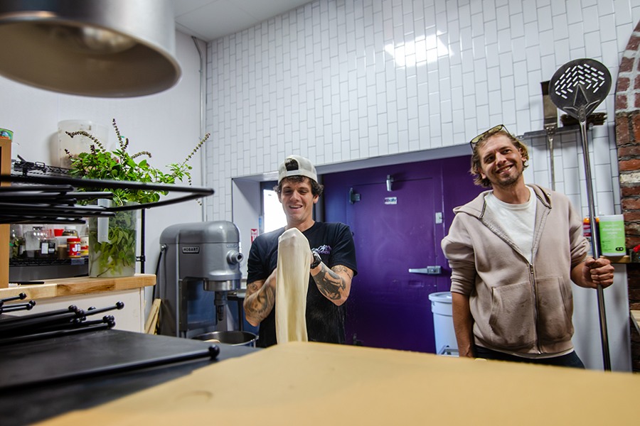 Two chefs stand in a kitchen, smiling. One is handling pizza dough while the other holds a long metal pizza peel.
