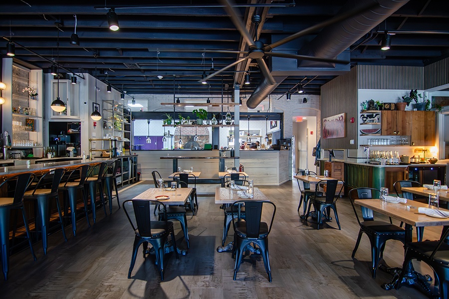 A restaurant interior without people in it. The vibe is neighborhood-y, with lots of light wood surfaces, pops of color, and plants.
