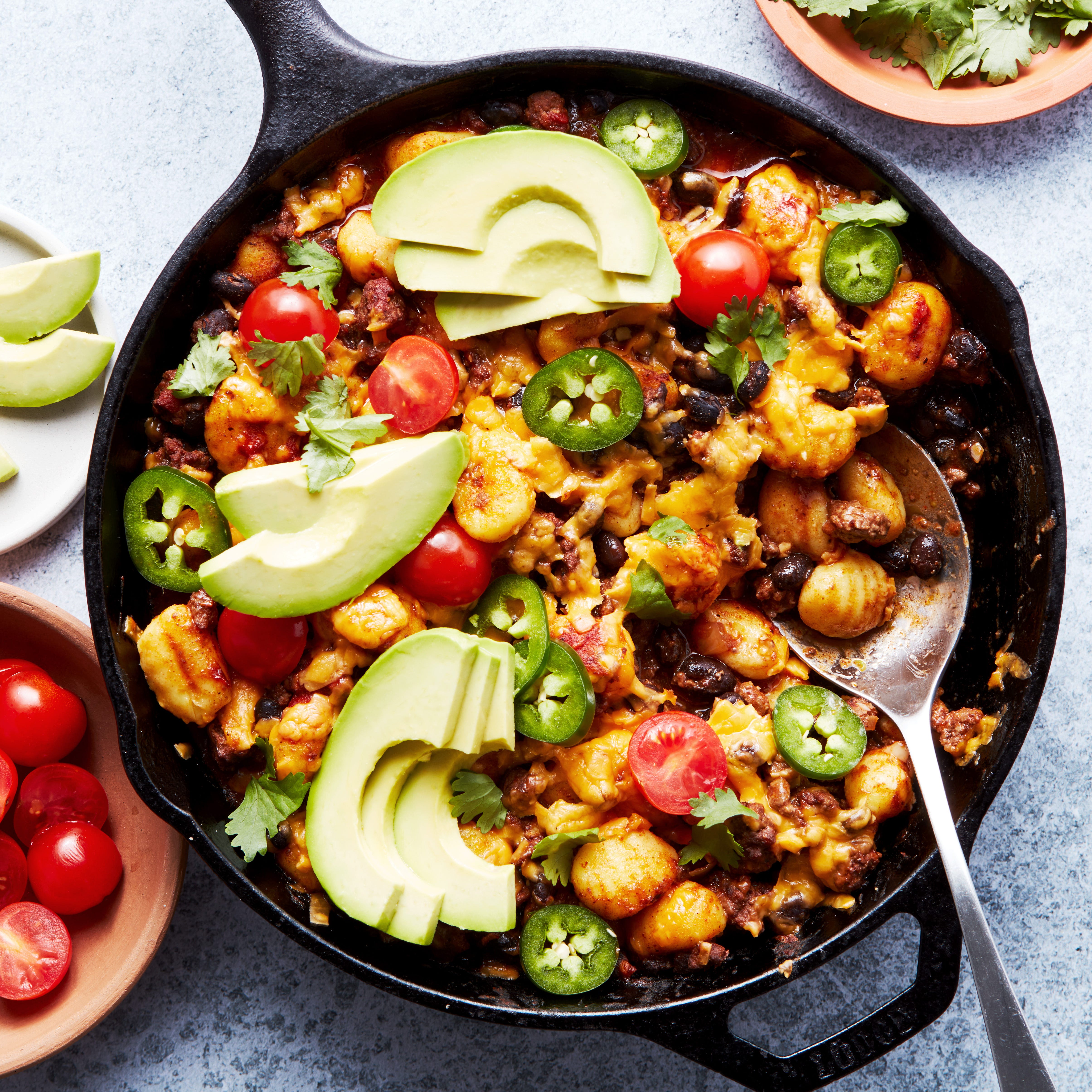 a pan with gnocchi covered in enchilada sauce, cheese, avocado, cilantro, and cherry tomatoes