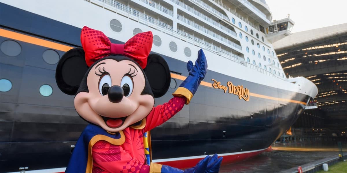 Minnie Mouse, dressed in a colorful outfit, poses and waves in front of a large cruise ship with the name "Disney Destiny" painted on its side. The ship is docked near a covered structure.