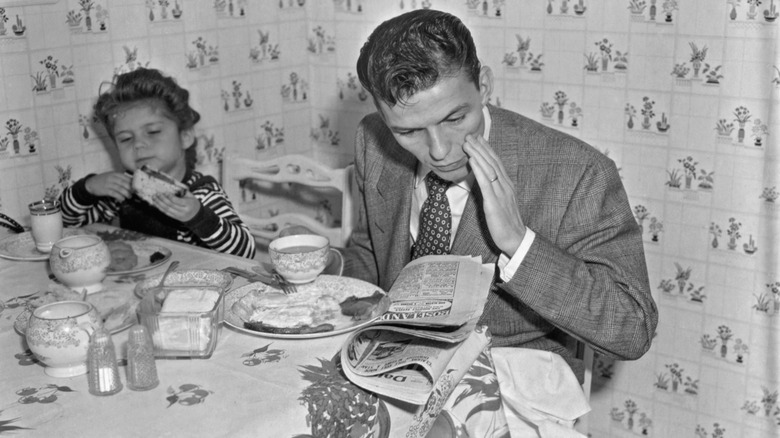 Black-and-white photo of Frank Sinatra looking down at a newspaper while eating breakfast