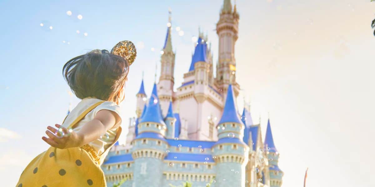 A young child wearing a yellow dress and Mickey Mouse ears reaches out joyfully toward a fairytale castle with blue rooftops under a bright, sunny sky.