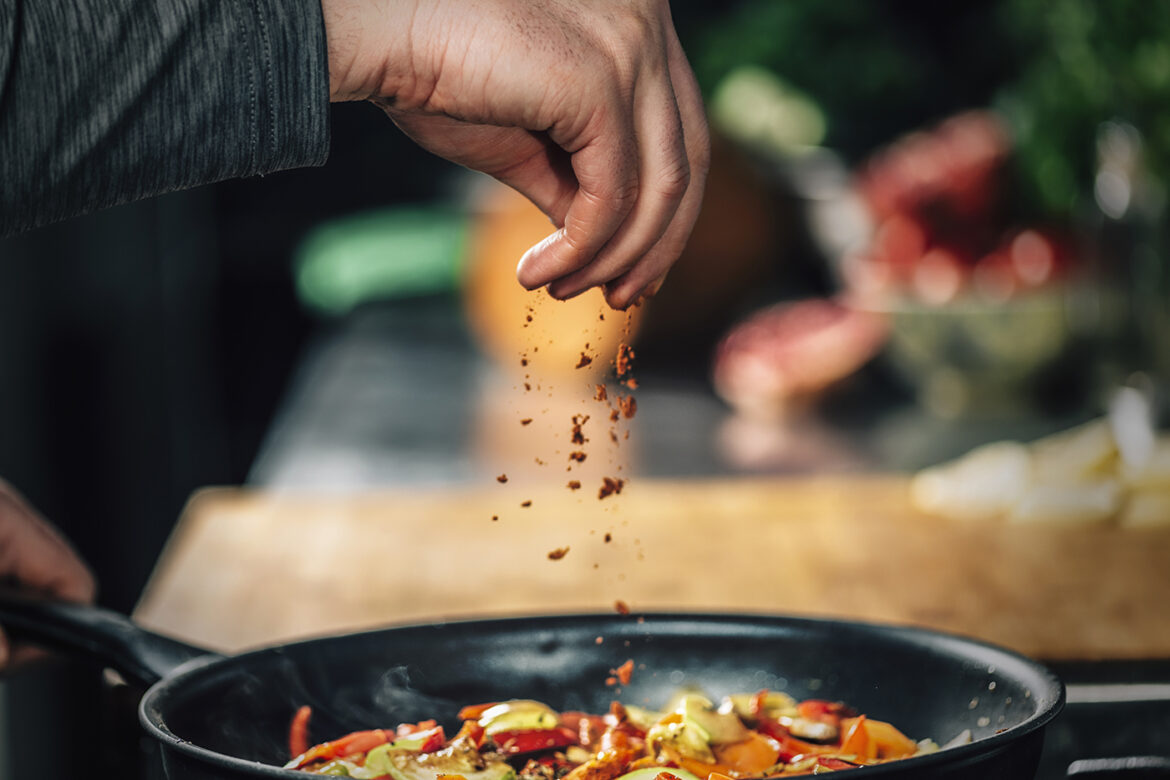 What Sets World Cuisines Apart? New Research Has the Answer A person's hand sprinkling chili flakes over a pan of food cooking on a stove.