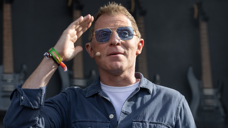 Bobby Flay with his hand up to his head wearing blue-tinted sunglasses and a face microphone