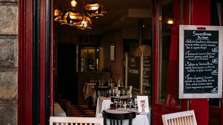 A glimpse into a classic French bistro, with a deep red door, tables draped with white linens and set with wine glasses, topped off with a sculptural lighting fixture
