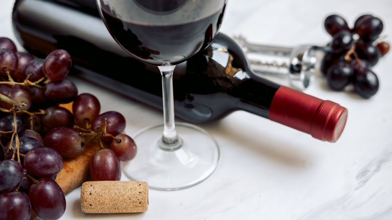 A bottle of red wine by a glass of wine, surrounded by grapes