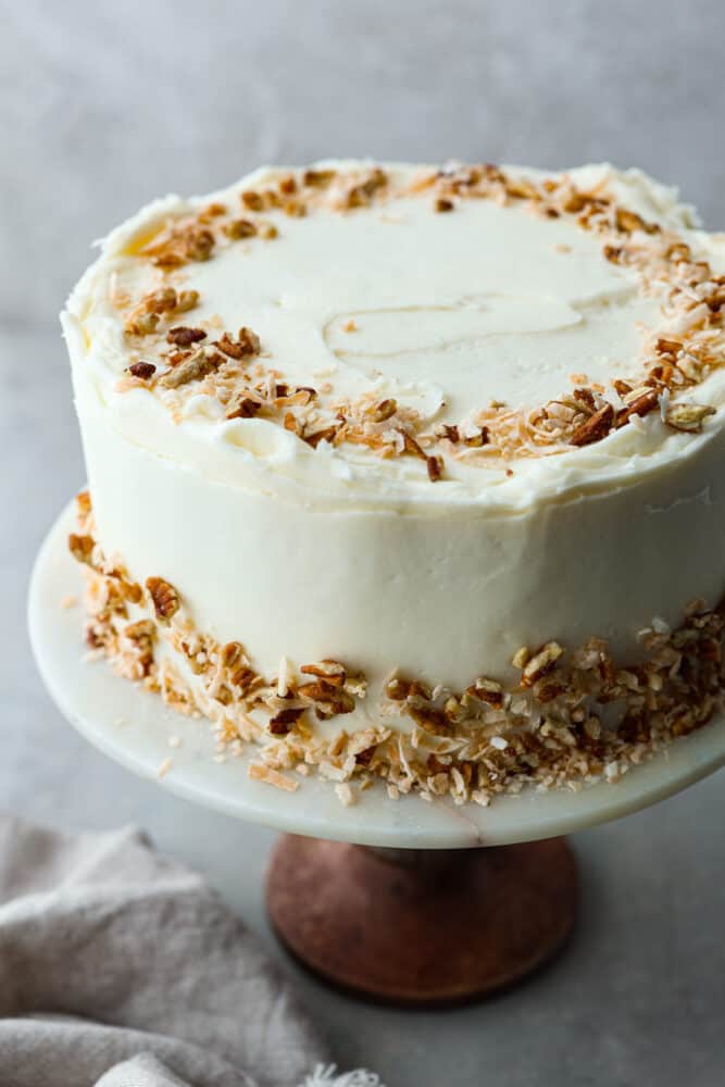 Take a bite of heaven with every forkful of this dreamy Italian cream cake. It's rich and nutty, covered with the most amazing cream cheese frosting! It's great for special occasions, but really, there's no wrong time to make it! A whole Italian cream cake on a stand topped with chopped pecans and coconut.