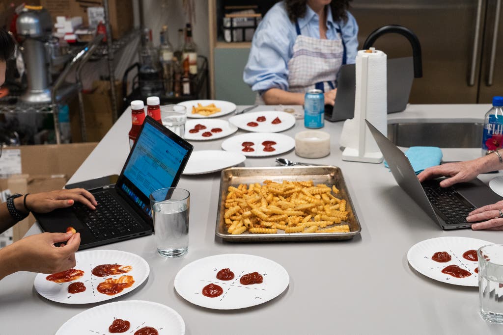 A few people in a kitchen typing on laptops and tasting French fries on the ketchup testing plates.
