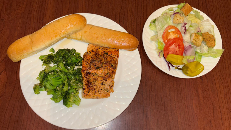 Plate with salmon, broccoli and breadsticks and plate with salad on wood table.