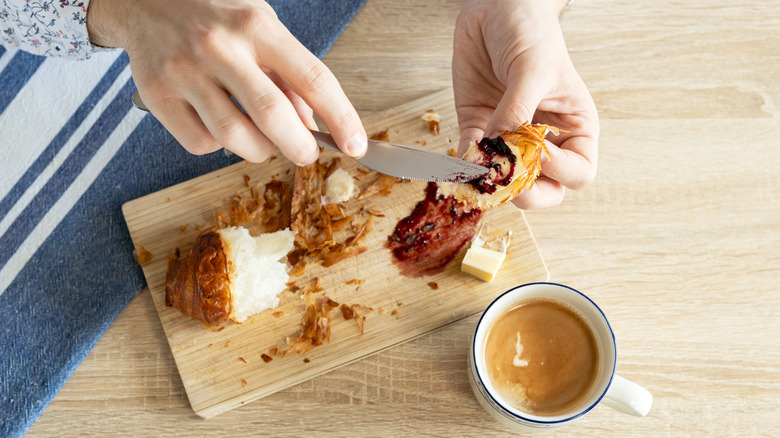 Two hands use a knife to spread jam across a torn croissant