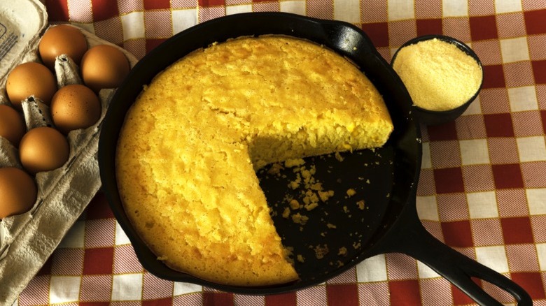 Cornbread in a cast iron skillet with a piece missing, alongside a carton of eggs on a red checkered tablecloth
