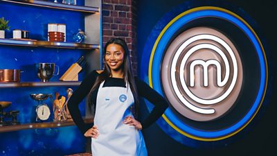 Uma wearing a white apron in the exposed brick MasterChef kitchen. Cooking equipment lines the shelves behind her and a large MasterChef logo in lights is to her left.