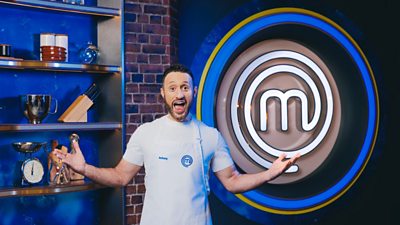 Antony Costa wearing a white apron in the exposed brick MasterChef kitchen. Cooking equipment lines the shelves behind him and a large MasterChef logo in lights is to his left.