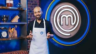 Gaz Choudhry wearing a white apron in the exposed brick MasterChef kitchen. Cooking equipment lines the shelves behind him and a large MasterChef logo in lights is to his left.