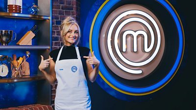 Katie wearing a white apron in the exposed brick MasterChef kitchen. Cooking equipment lines the shelves behind her and a large MasterChef logo in lights is to her left.