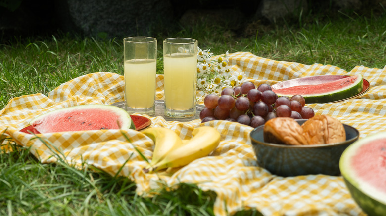 Two glasses of lemonade and fresh fruits on checkered yellow and white picnic blanket.