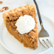 one slice of praline pumpkin pie with pecan streusel topping and a dollop of whipped cream on a white plate with a black handled fork