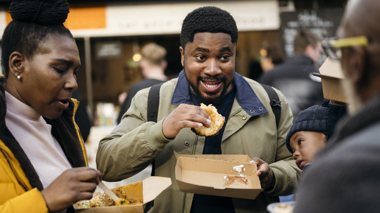 Waist-up view of smiling young Black man with baby boy and grandparents standing outdoors talking and eating specialty food in Southwark