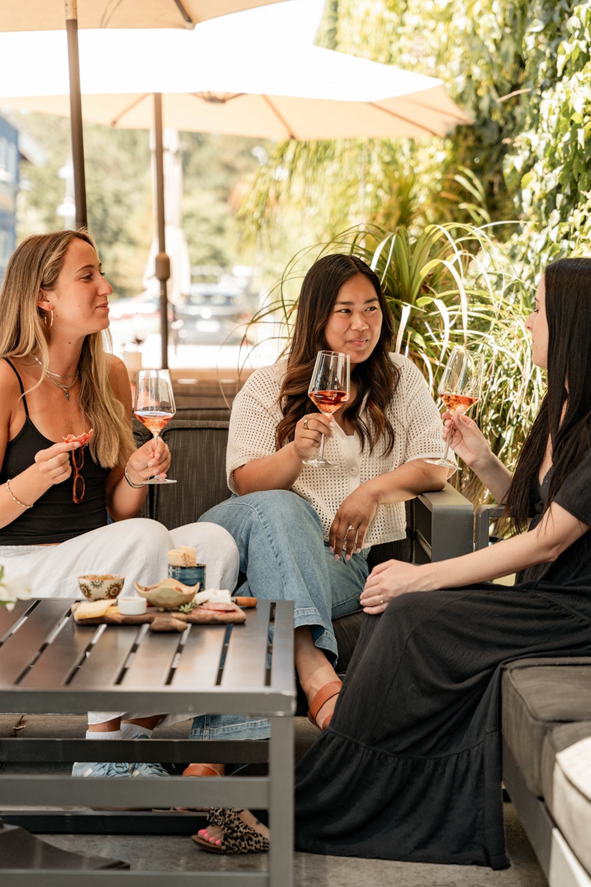 Three women enjoying wine on a patio with sun-shading umbrellas next to an overgrown wall.