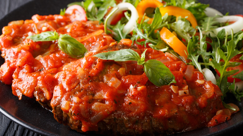 Black plate with steak covered with tomato sauce and salad