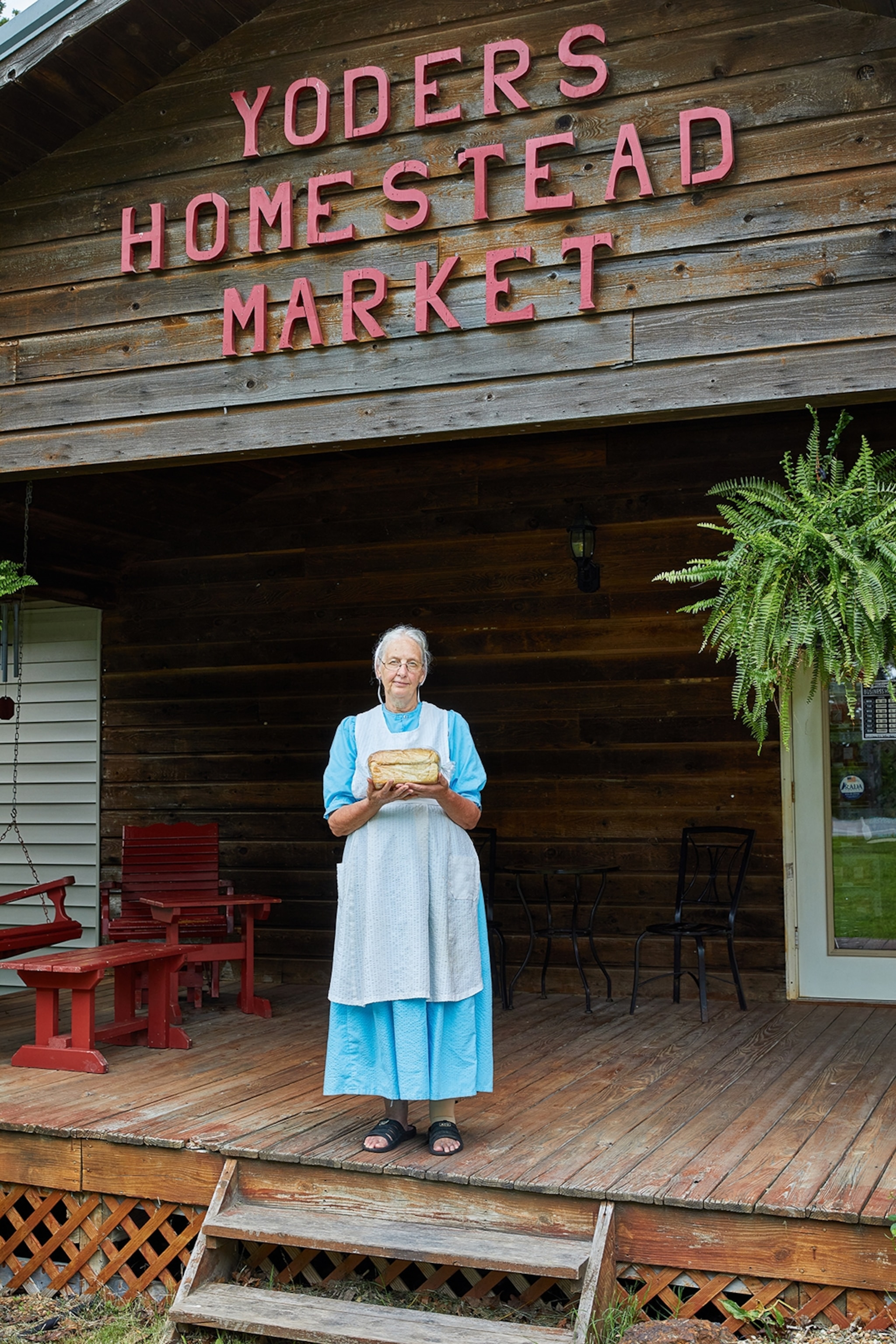 An eldery woman, wearing a long Amish dress and bonnet, on a wooden porch, holding out a pie.