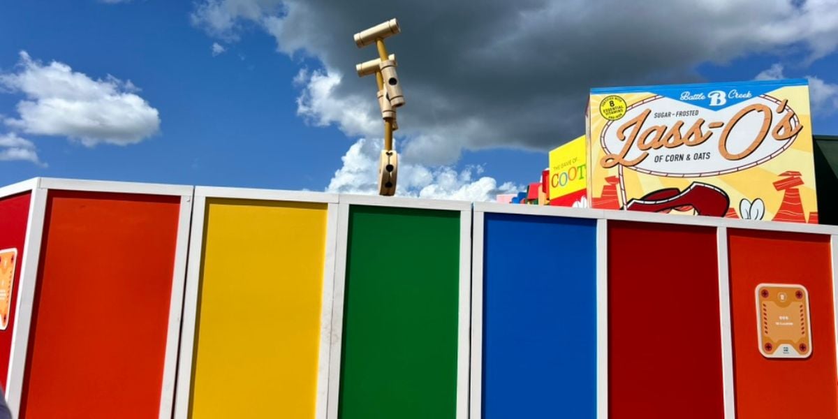 A colorful fence with red, yellow, green, and blue panels stands under a partly cloudy sky. Behind it is a large cereal box labeled "Lassi O's" and a wooden periscope structure, evoking the playful charm of a Disney-inspired scene.