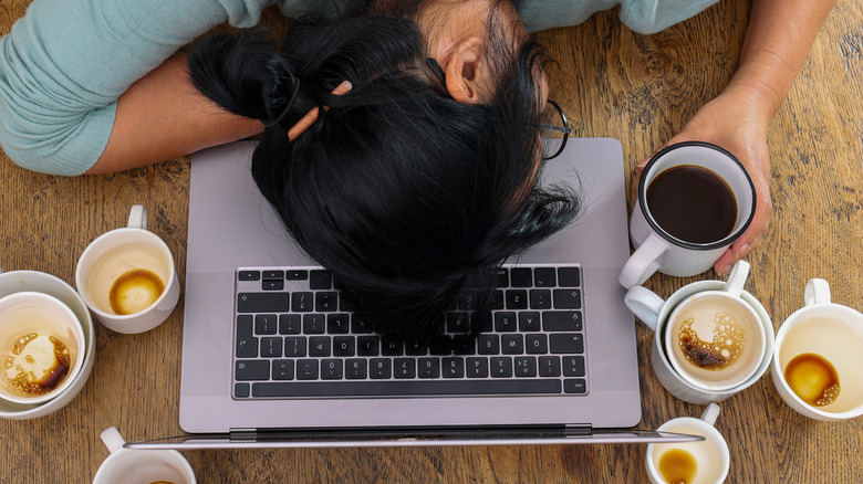 A woman asleep at a computer surrounded by piles of coffee cups.