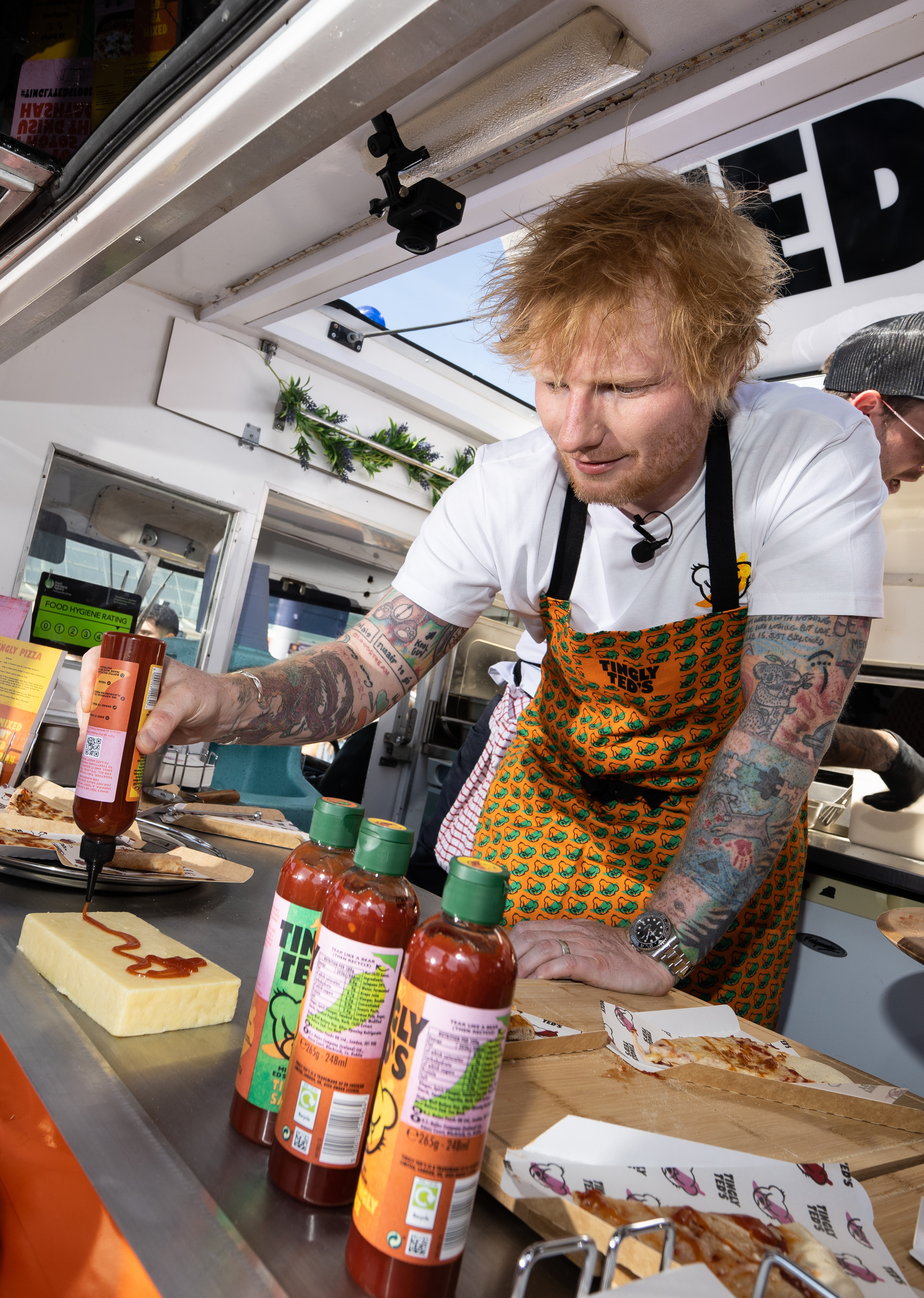 Ed Sheeran promoting his Tingly Ted's hot sauce brand by squeezing sauce onto a block of cheese.