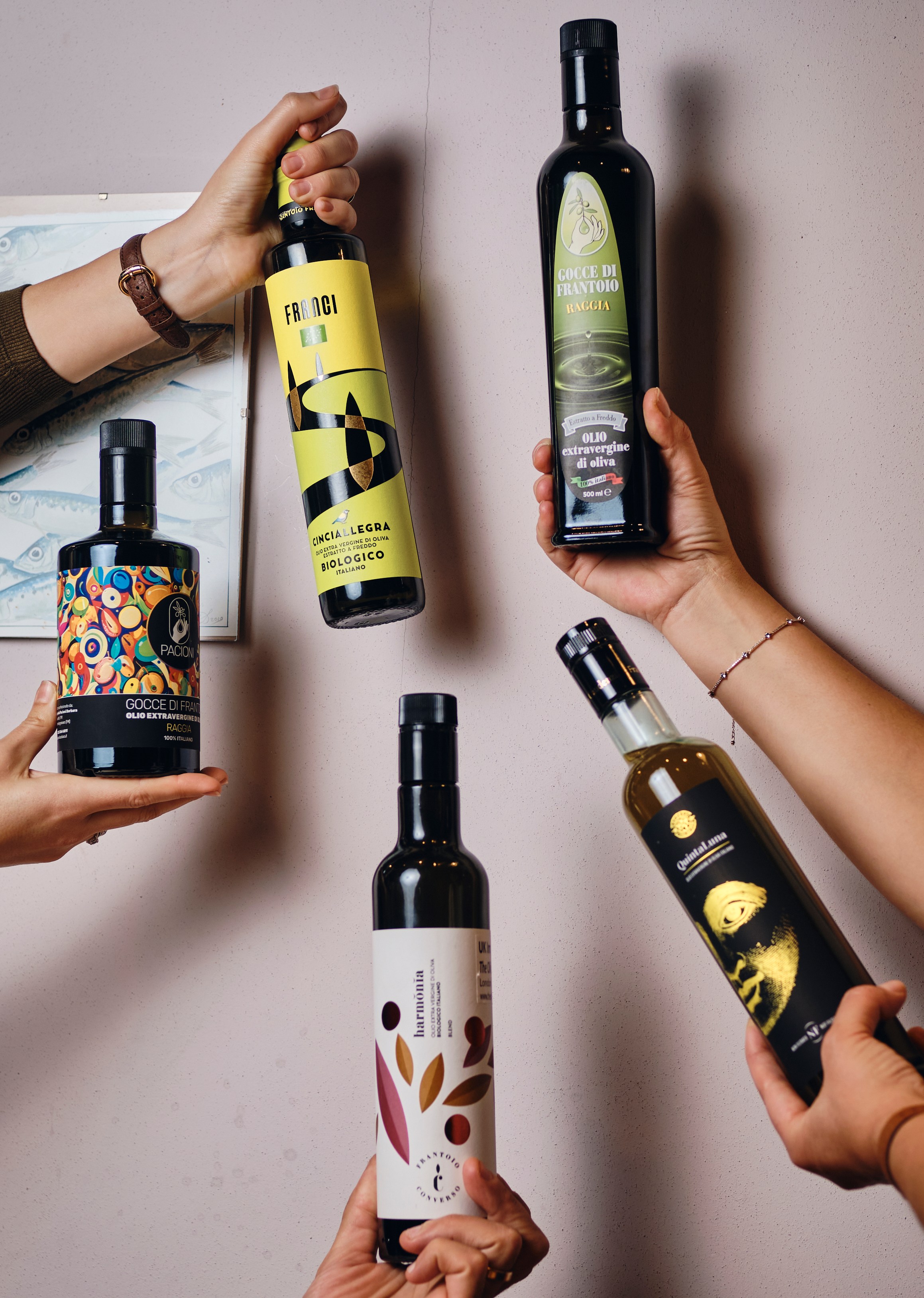Hands holding four bottles of olive oil with different labels against a light pink wall.