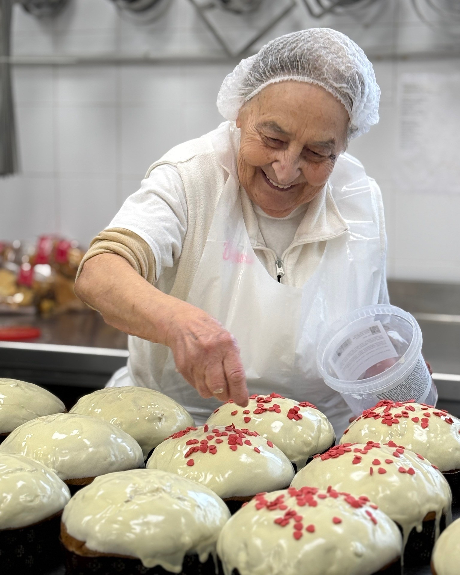 An elderly woman with a hairnet and apron smiles while sprinkling red heart-shaped decorations onto white-frosted cakes.