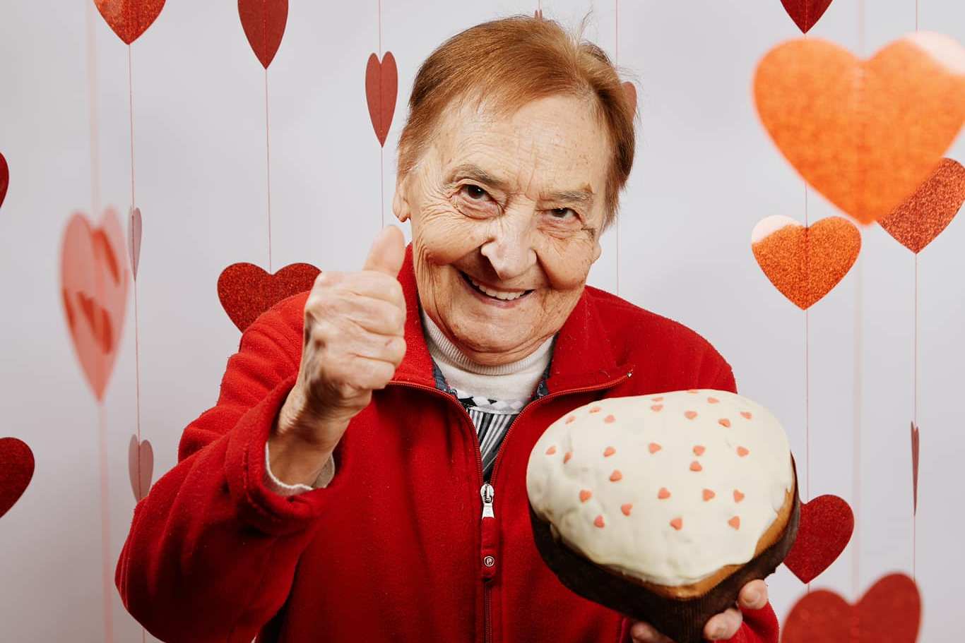 Nonna Silvi giving a thumbs up while holding a cake with a white topping and small heart sprinkles.