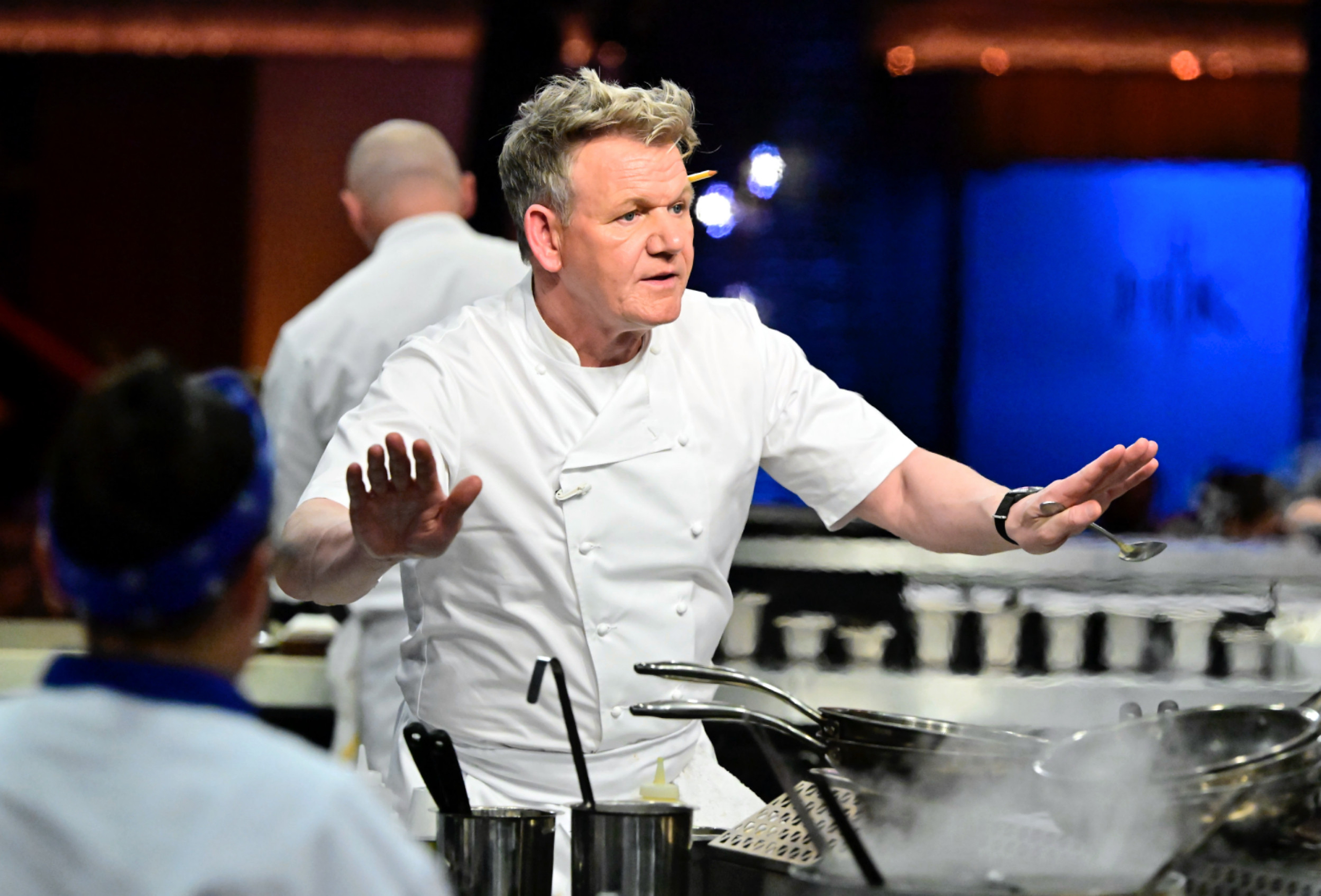 Gordon Ramsay, in a white chef's coat, gestures with both hands in a kitchen, holding a spoon in one.