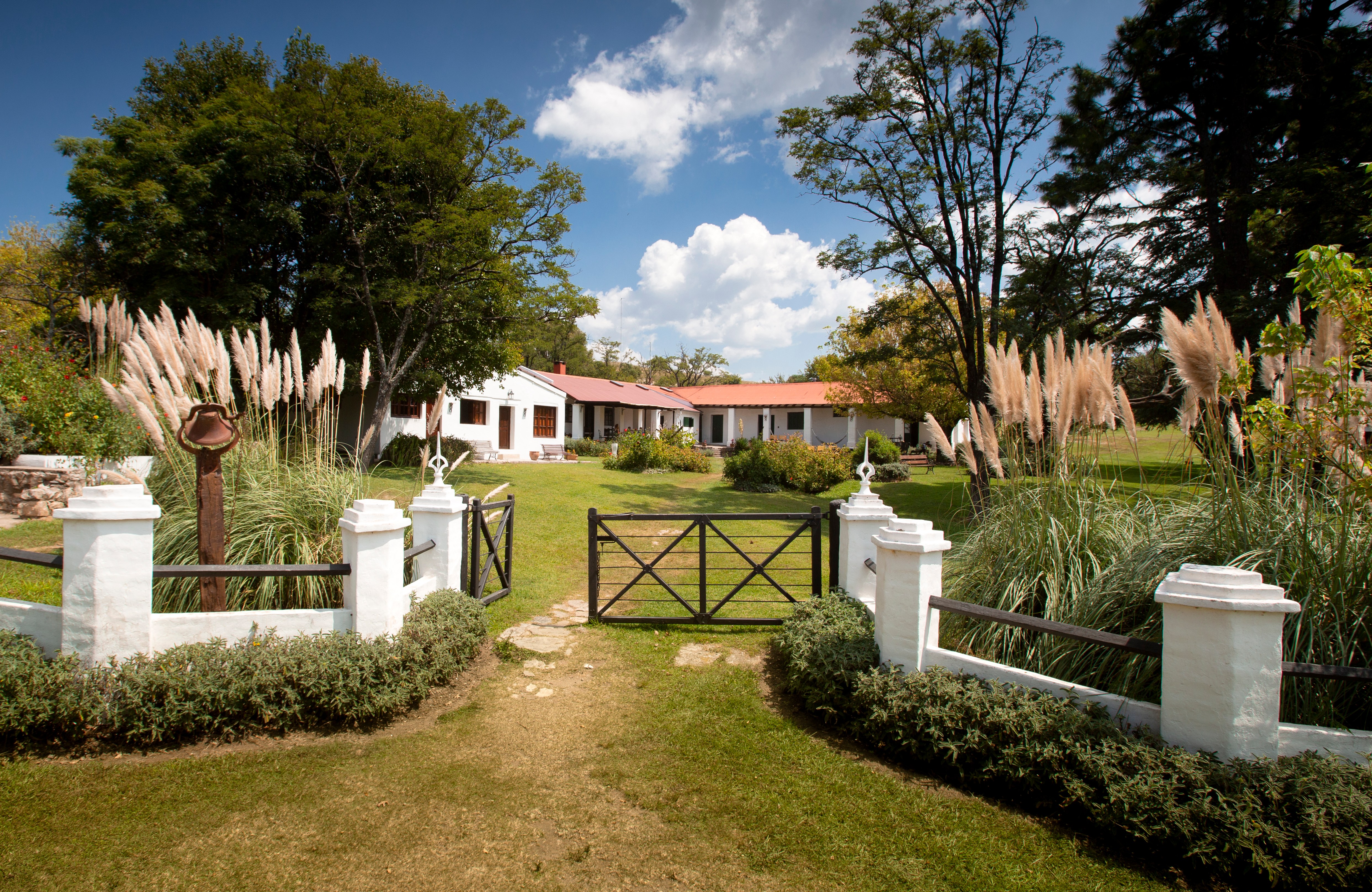 A white house with a red roof seen through a black gate, surrounded by trees and tall grass, under a blue sky with white clouds.