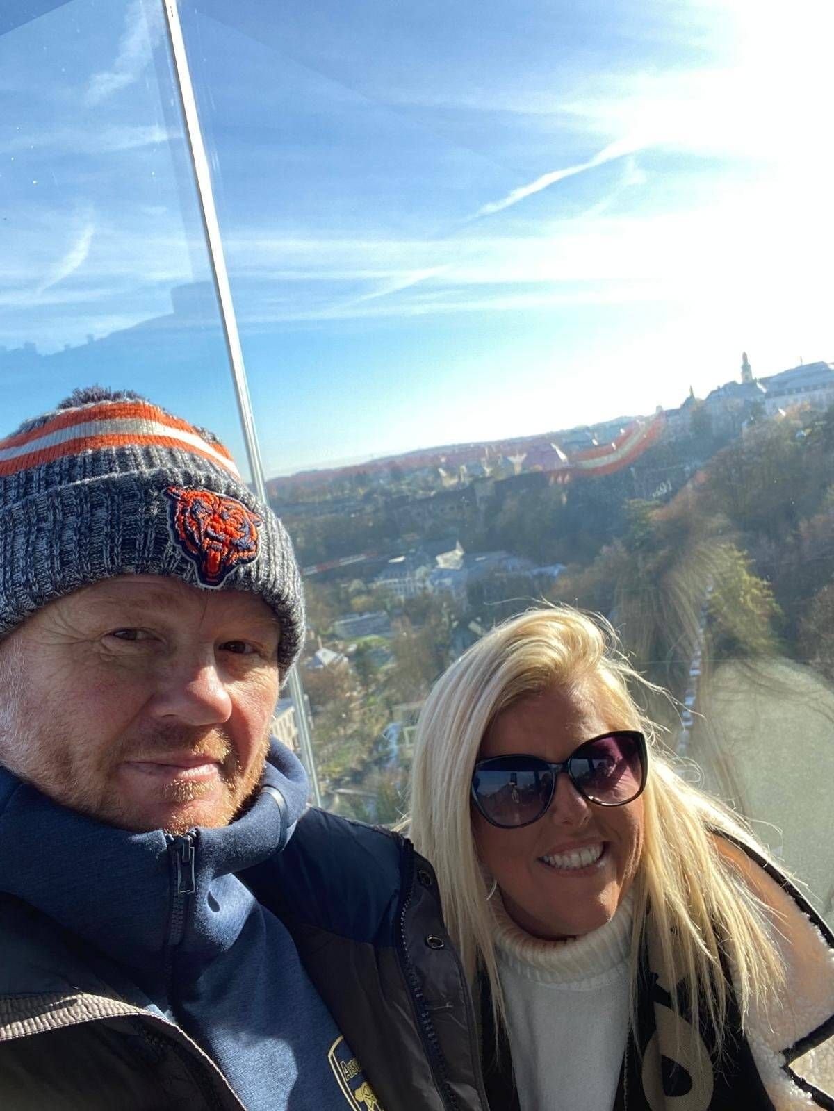 Two individuals are enjoying a scenic view from a high vantage point, dressed in winter attire. The male figure sports a beanie and a jacket, while the female figure dons sunglasses and a white top. They appear to be in a glass-enclosed observation area, with a mountainous landscape and clear blue sky visible in the background.