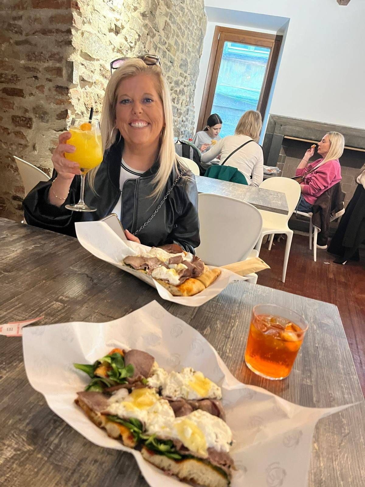 A woman is seated at a table in a dining area, holding a glass of orange juice. The table has two plates with various foods, including what appears to be a sandwich and a beverage. In the background, there are other individuals seated at different tables, with a brick wall and a window visible.