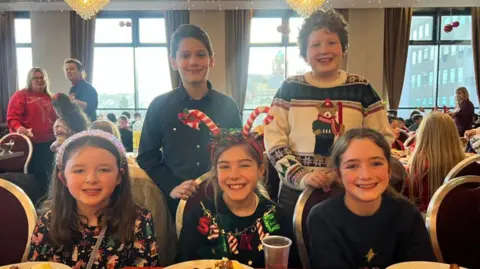 Five children smiling at the camera. At the back there are two boys and in the front three girls. All of them are wearing Christmas themed outfits. Behind them are windows. 