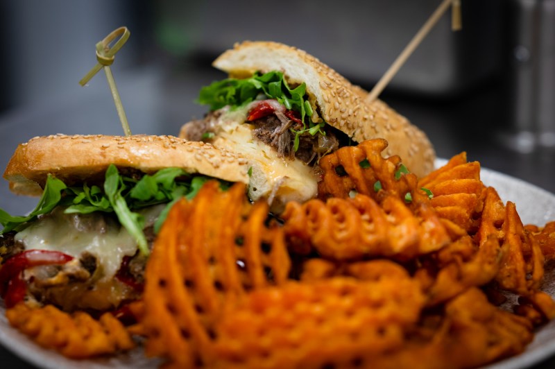 A stacked sandwich on a sesame seed bun filled with meat, melted cheese, and fresh arugula, served alongside seasoned wavy-cut sweet potato fries on a white plate.