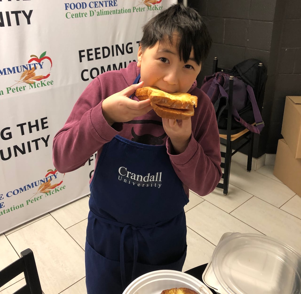A boy bites into a peanut butter and jelly french toast with his hands. He's wearing a blue apron. 