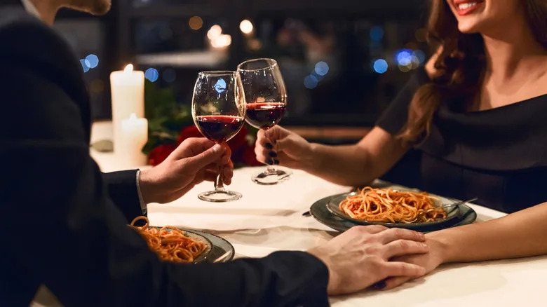 A couple toasting wine glasses over plates of spaghetti at a restaurant
