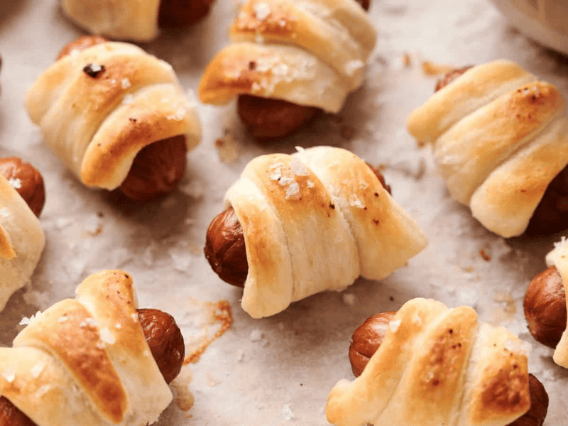 Pigs in a Blanket, small sausages wrapped in puff pastry, baked until golden brown, and sprinkled with coarse salt are displayed on a baking sheet lined with parchment paper.