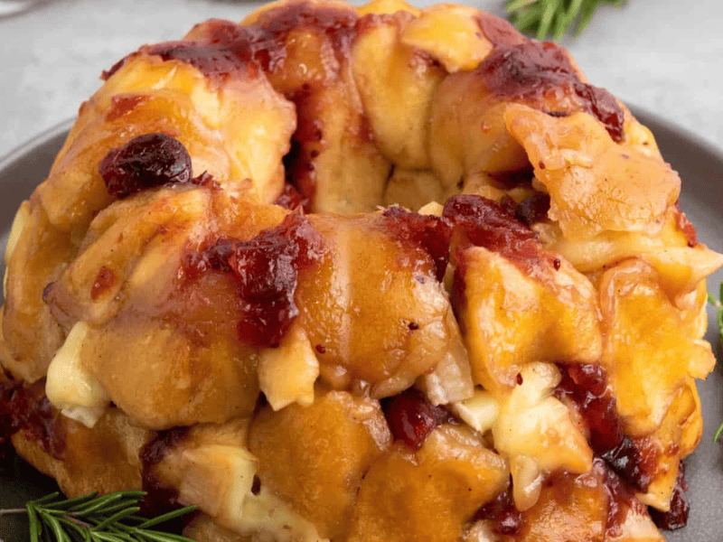 A close-up of a bundt-shaped bread loaf with visible chunks of cheese and cranberries, garnished with rosemary on the side.