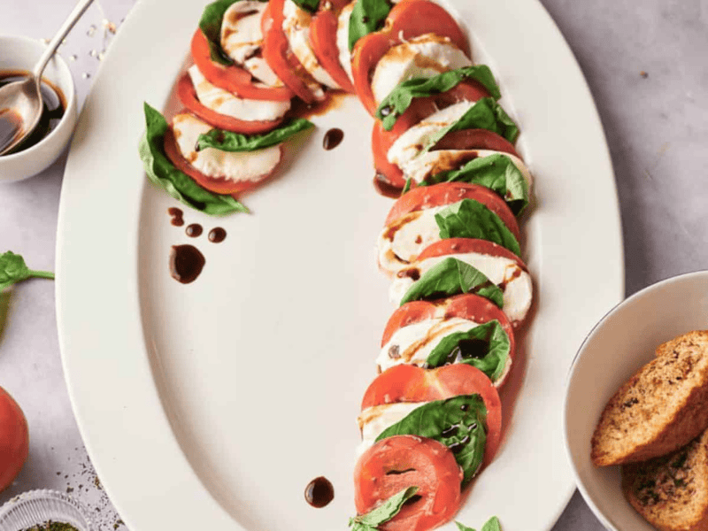 Sliced tomatoes, mozzarella, and basil arranged in a curve on a white platter, drizzled with balsamic glaze. A bowl of crostini and balsamic are on the side.