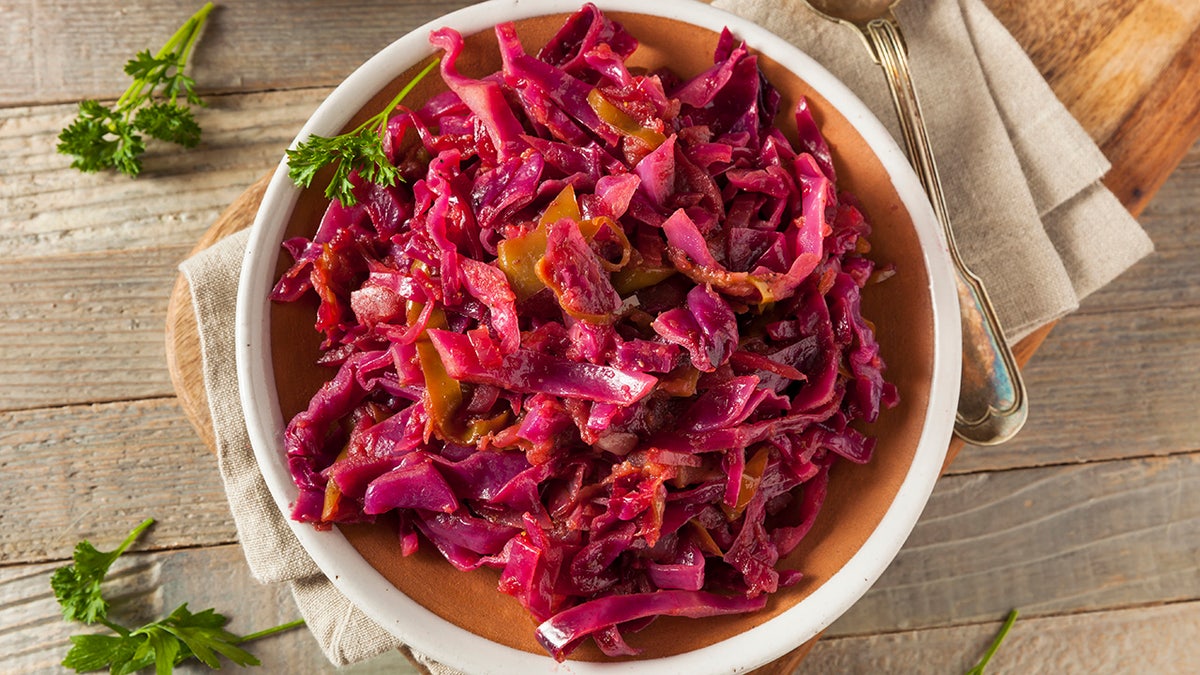 Bowl of braised red cabbage on wooden table, sprinkled with green garnish, spoon and napkin beside bowl.