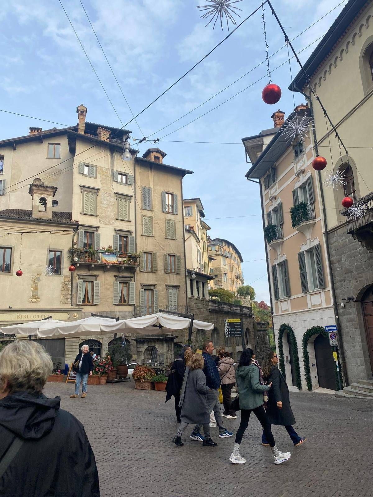 An urban street scene with pedestrians walking past buildings with balconies and awnings, under a bright blue sky with hanging lanterns.